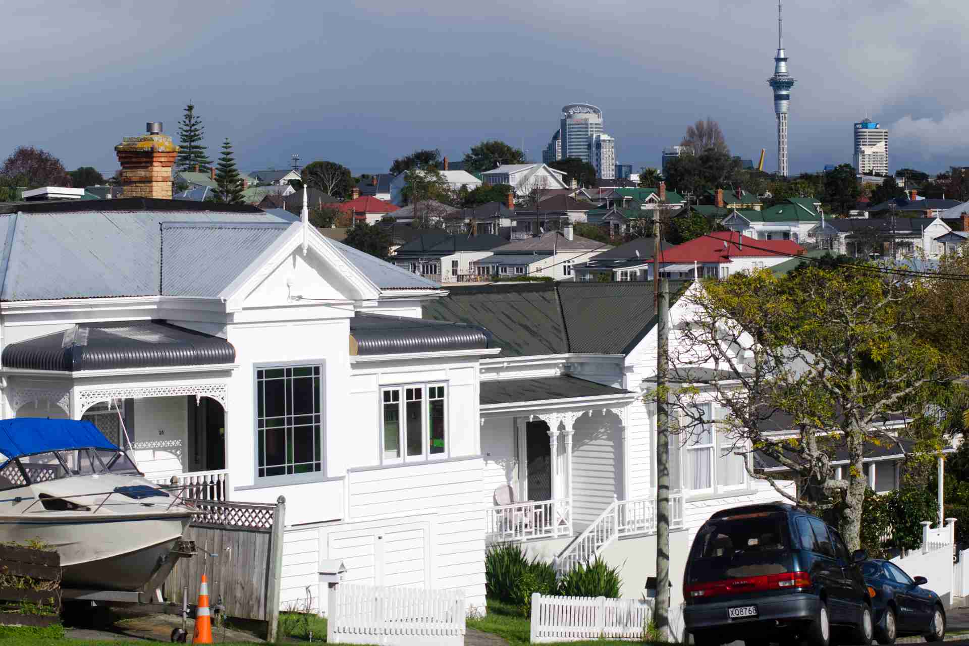 Residential street with white houses, parked cars, and the Sky Tower in the background.