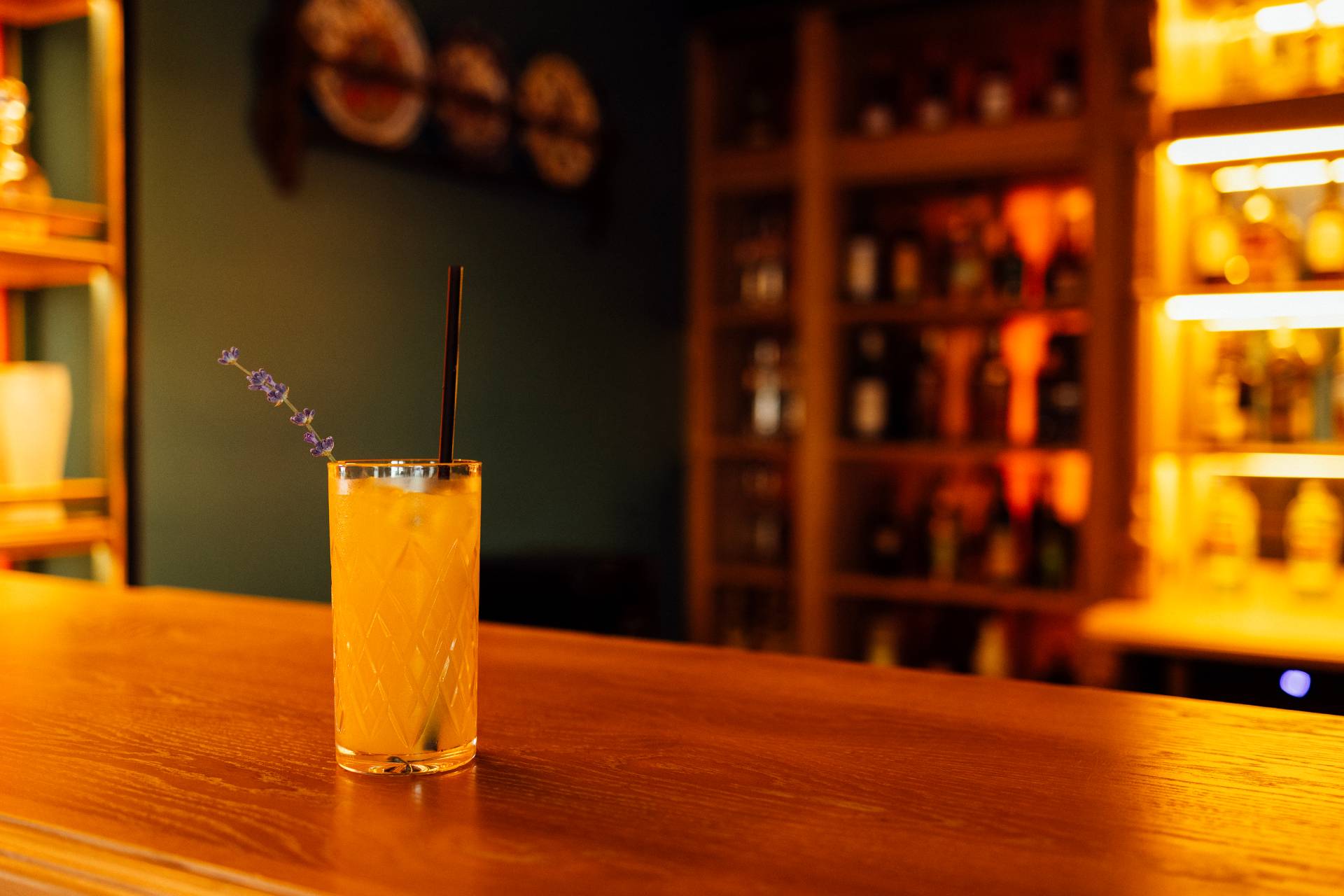 A tall glass of orange cocktail with ice and a lavender sprig garnish sits on a wooden bar counter in a warmly lit bar