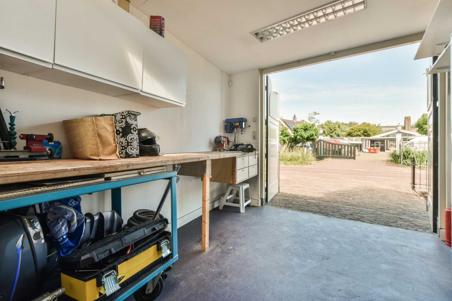 A bright garage workshop with a wooden workbench, tools, and storage shelves, opening out to a sunny driveway.