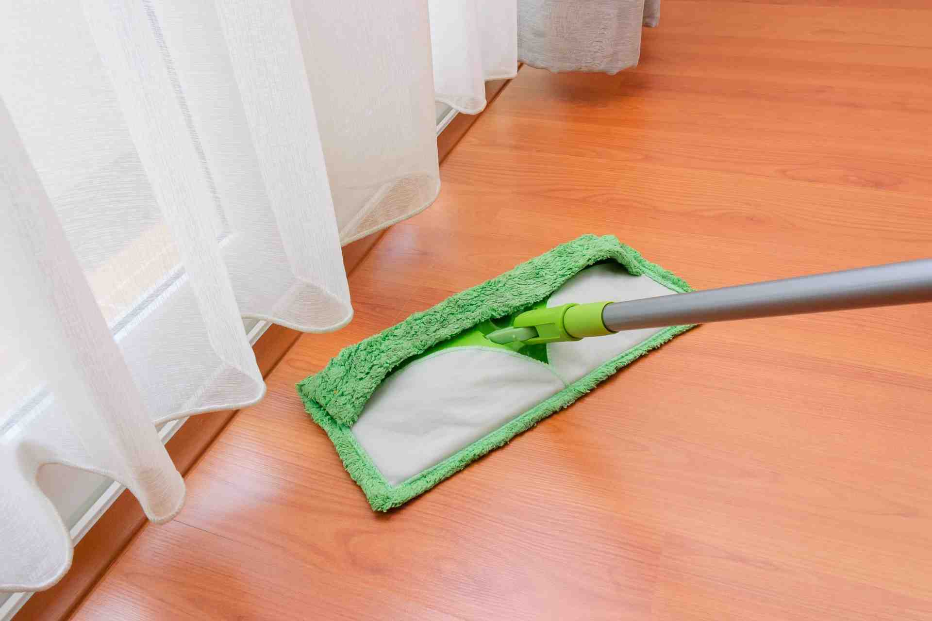A green flat mop cleaning a wooden floor near white curtains