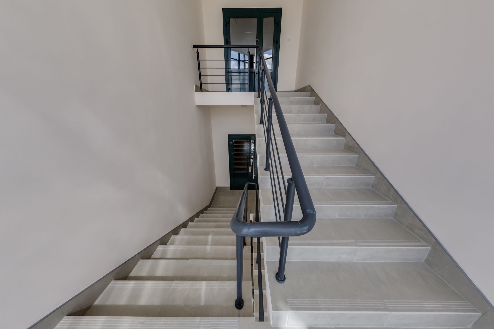 Indoor stairwell with light tiled steps, metal railings, and doors at the top and bottom landings