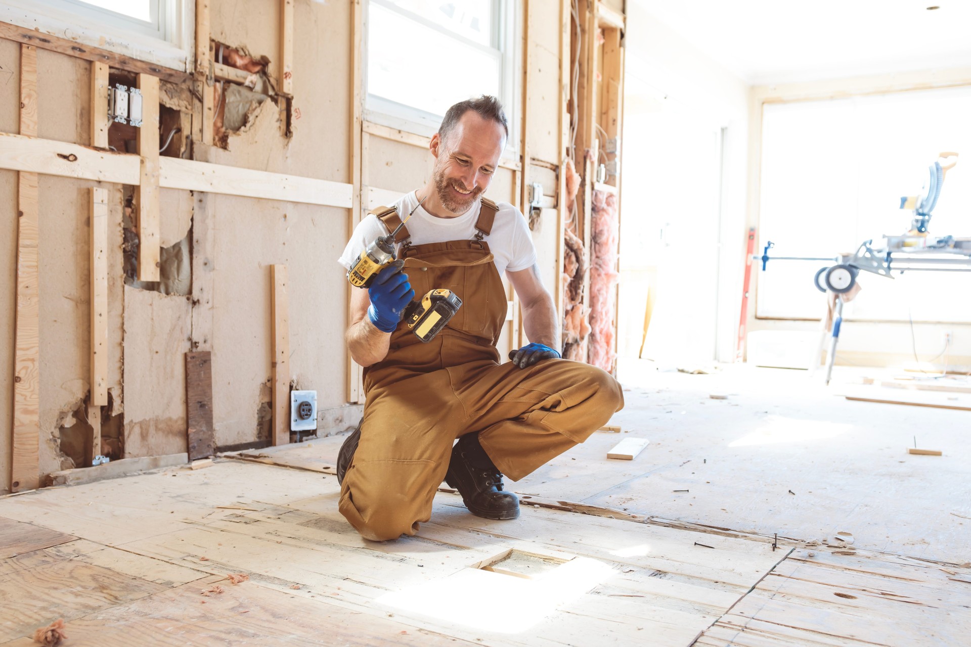 Man kneeling on a subfloor during home renovation, smiling while holding a cordless drill.