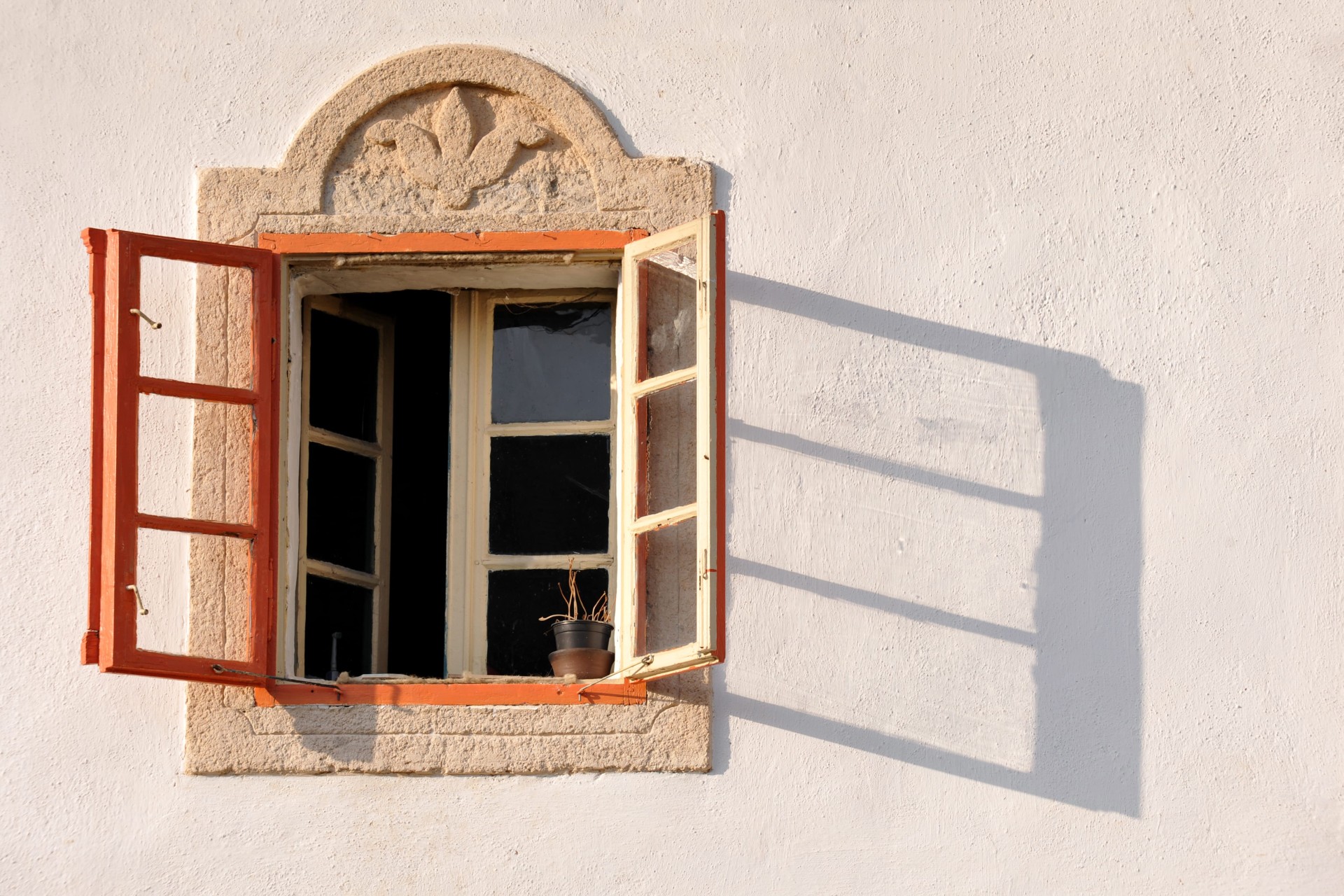 Open wooden window with red shutters casting a long shadow on a white wall