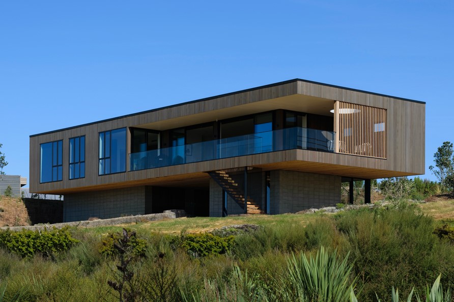 A modern rectangular house elevated on a concrete base with large glass windows, a balcony, and wooden exterior panels on a grassy hillside.