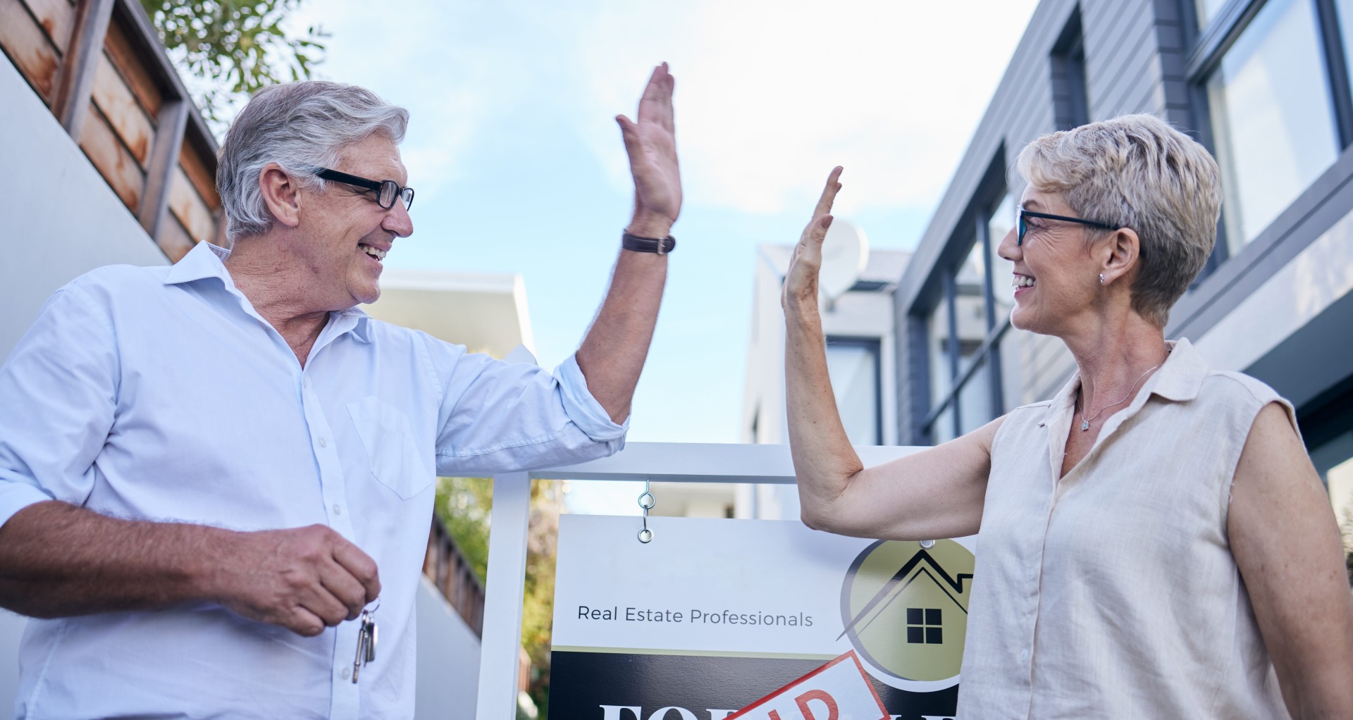 Older couple high-fiving outside a house with a sold real estate sign, celebrating a property sale