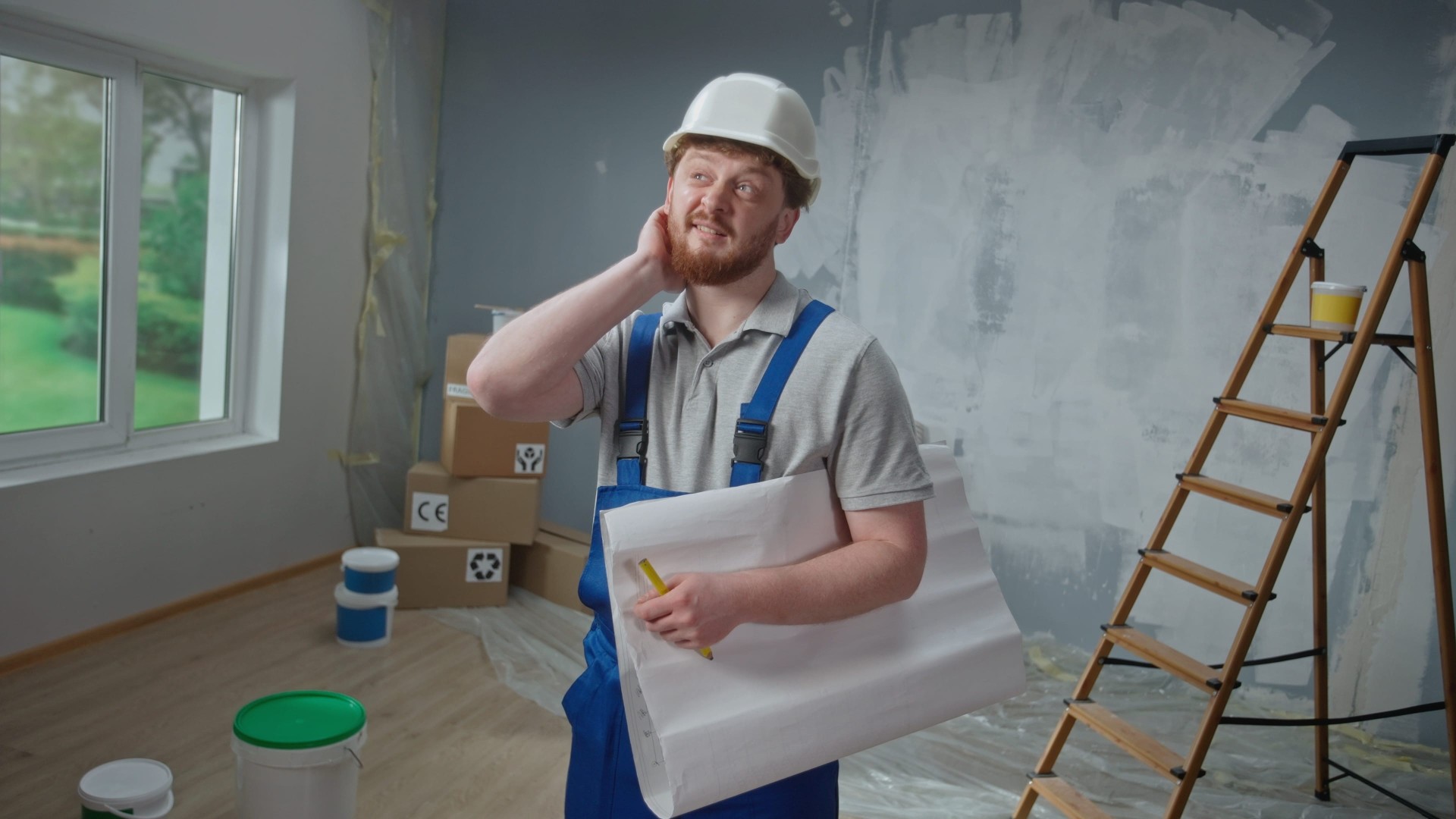 A construction worker in a partially renovated room holding blueprints and looking thoughtful beside a ladder and paint supplies