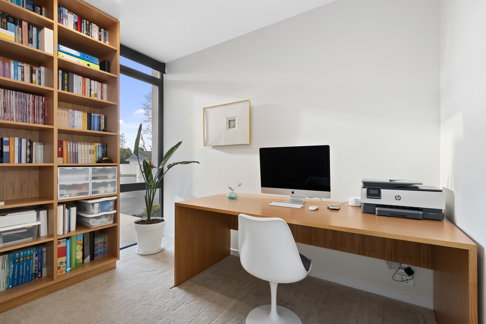 Bright home office with a timber desk, white chair, built-in bookshelves, printer and tall window beside an indoor plant