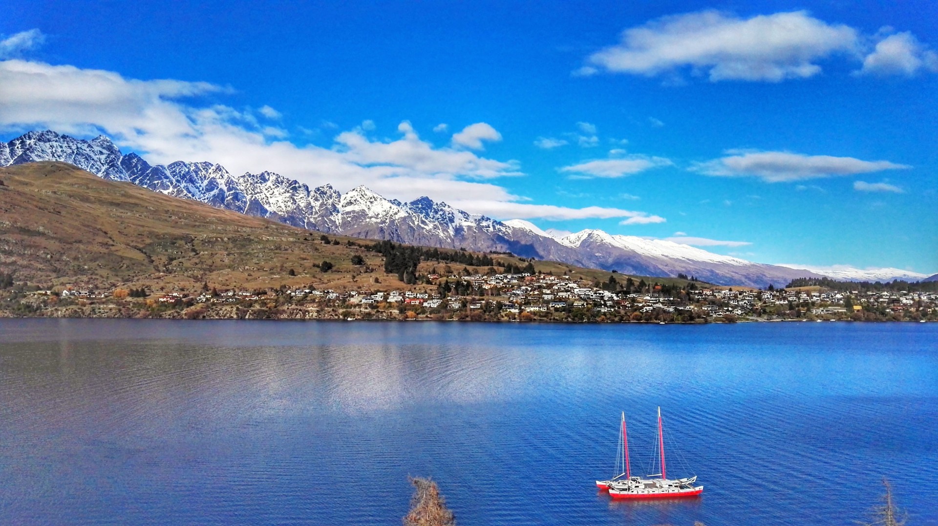 Snow-capped mountains overlooking a calm blue lake with a red sailboat in the foreground