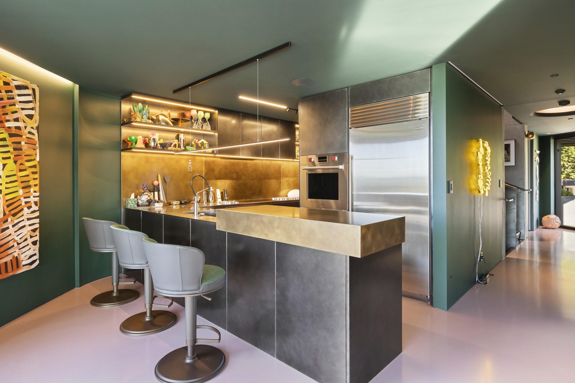 A modern kitchen with green walls, pink flooring, metallic cabinetry, bar stools, and warm shelf lighting