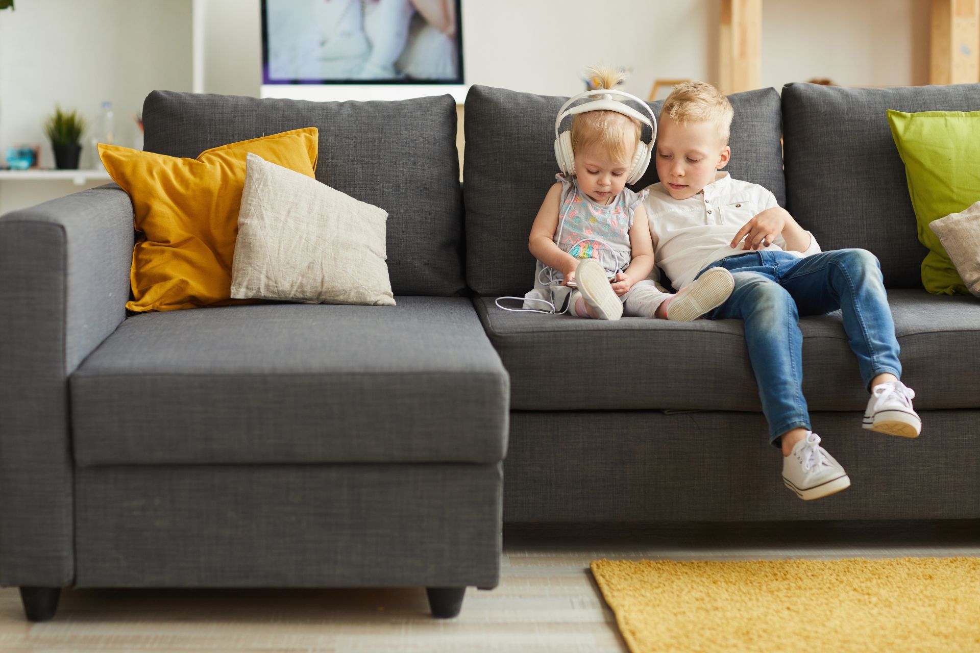 Toddler and young child relaxing together on an L-shaped sofa in a bright living room.