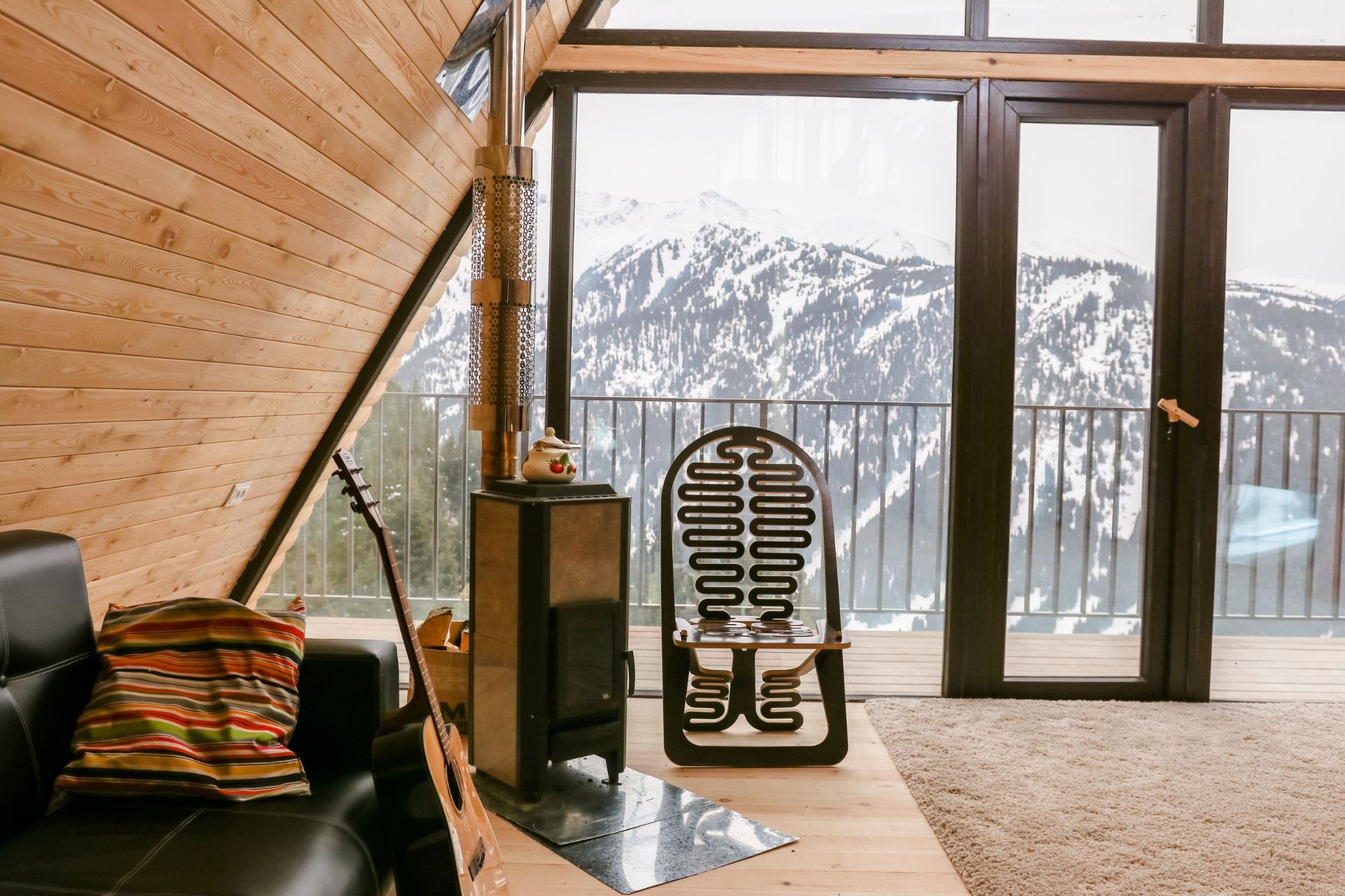 Alpine-style living room with timber walls, a wood-burning stove, leather sofa and snowy mountain views through large windows.