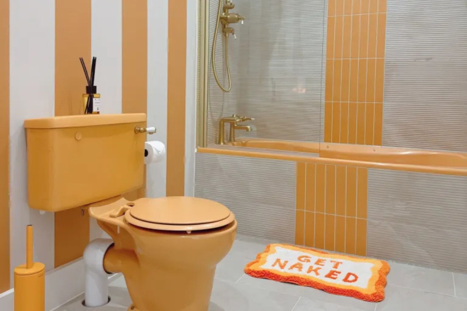 Orange bathroom featuring a mustard-yellow toilet and cistern, striped walls, grey shower tiles and a matching orange bath mat.