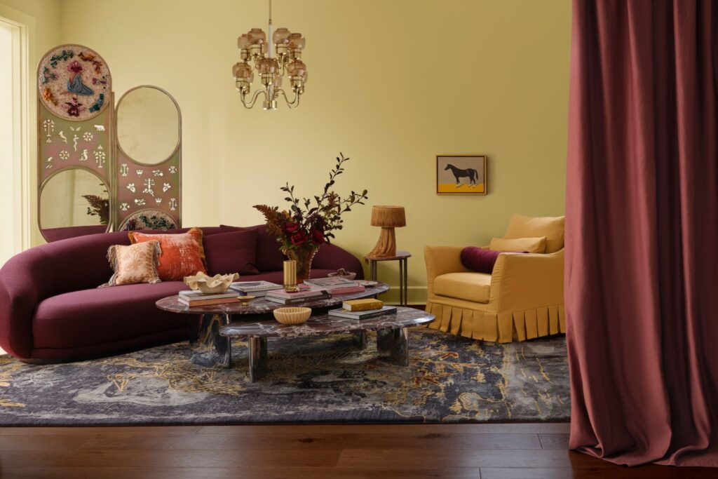 Jewel-toned living room featuring a deep burgundy sofa, mustard yellow armchair, patterned rug and rich plum curtains against warm yellow walls.