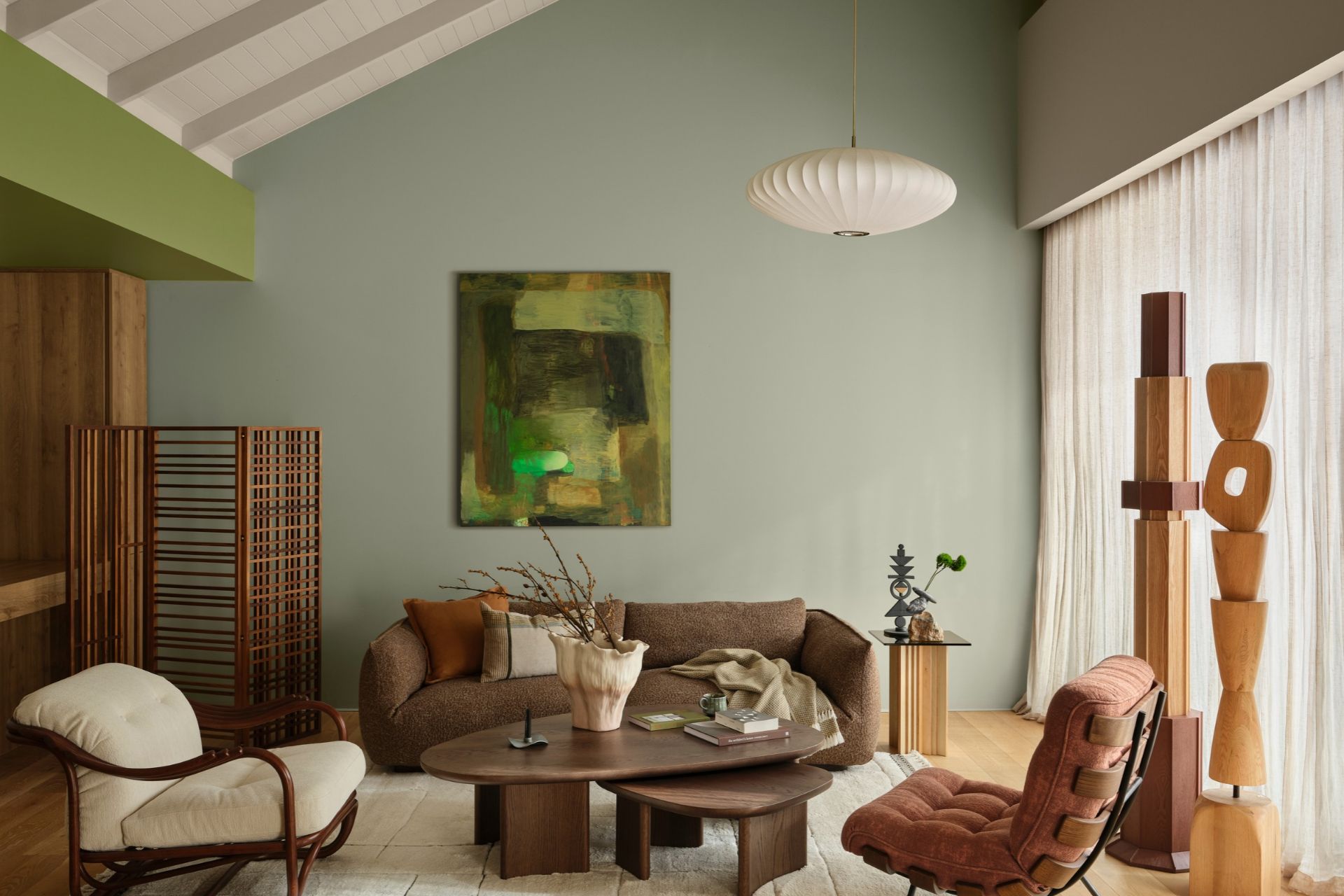 Modern living room featuring muted green walls, warm wooden furniture, textured upholstery, sculptural décor and soft natural lighting.
